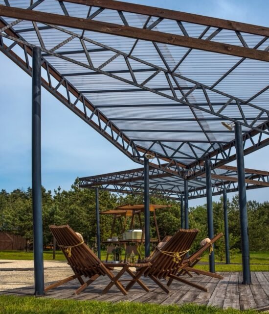 A stylish gazebo with a metal frame and a transparent roof creates the perfect place for outdoor recreation. Wooden deck chairs and a green landscape place to relax