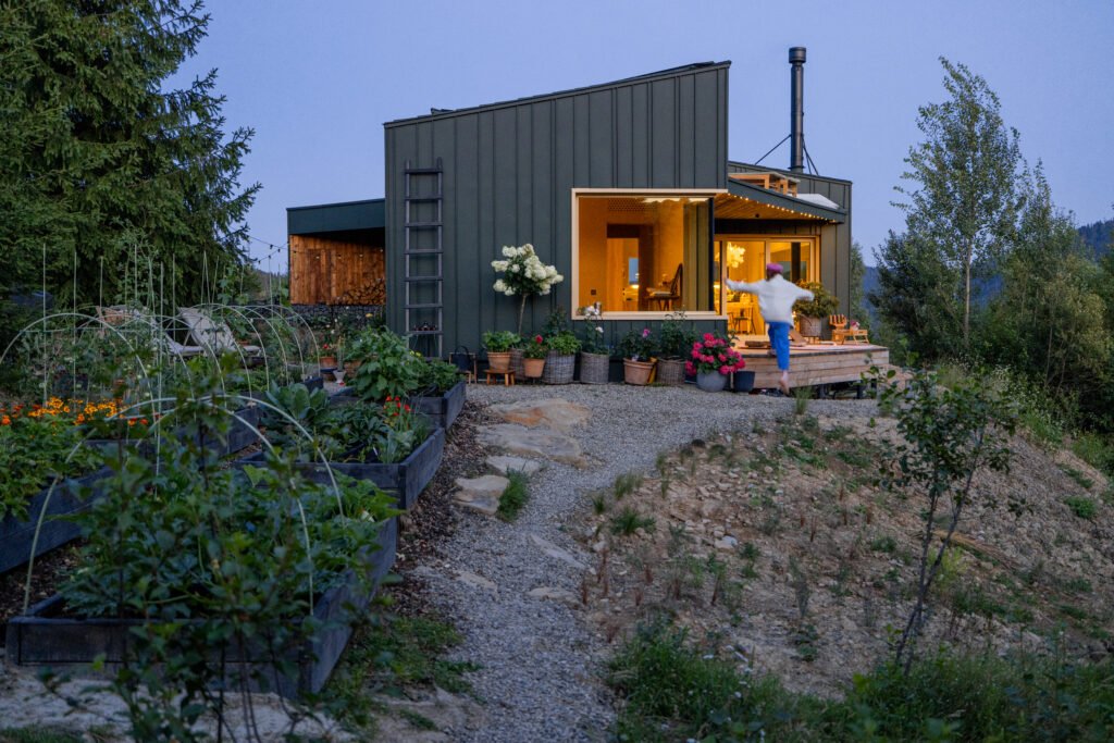 Modern cabin at dusk with soft, warm interior lighting illuminating the cozy living space. Beautiful garden in bloom complements the tranquility of the mountain surroundings